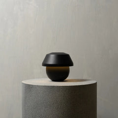 A modern black mushroom-shaped table lamp with a spherical base, displayed on a textured gray cylindrical pedestal against a neutral gray wall.