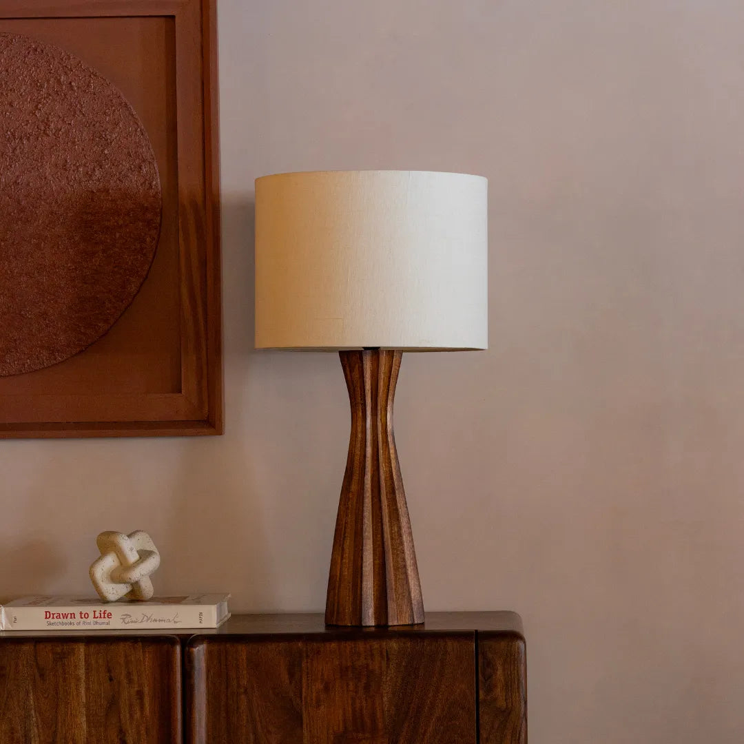 Wooden Tern Table Lamp with a white shade & wooden stand on a wooden surface against a beige wall.