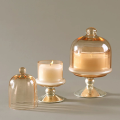 Sundance Aura Candle - amber glass cloche domes and candle holders on a neutral gray background, including one open dome, one candle in a glass holder, and one larger candle under a cloche.