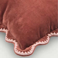 A close-up of a plush, rust-brown velvet throw pillow with a scalloped edge trimmed in white and rust-brown fabric.