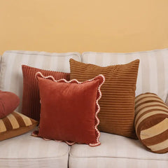 A set of decorative throw pillows arranged on a white sofa against a warm beige wall. The collection includes rust red velvet pillows with scalloped edges, a brown corduroy pillow, striped pillows, and a round bolster pillow.