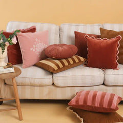 A styled collection of decorative throw pillows in various colors and textures arranged on a cream-colored sofa against a warm beige wall, with a small wooden side table on the left holding a vase of flowers and books.