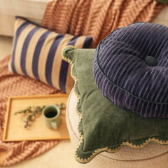 A styled collection of home decor pillows, featuring a round tufted navy corduroy cushion on top of a green scalloped velvet pillow, with a blue and beige striped pillow in the background on a patterned throw blanket.