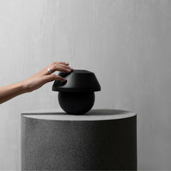 A matte black modern speaker with a rounded dome top sits on a textured gray cylindrical pedestal, with a hand reaching toward the top of the speaker against a light gray wall background.
