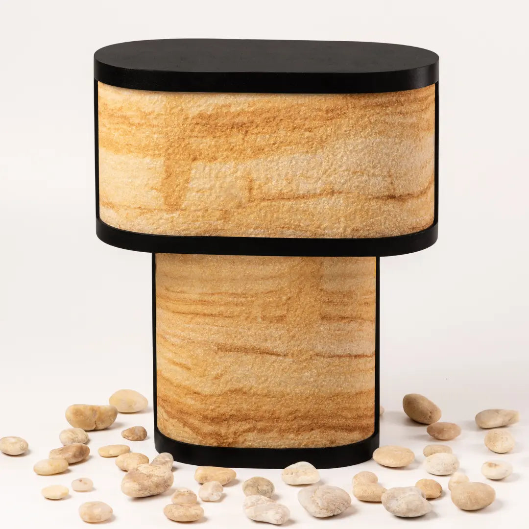 A cylindrical Niko table lamp with a teak wood sandstone base and a black powder-coated mild steel top, surrounded by scattered stones.