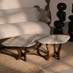 Two nested oval coffee tables with polished light gray and white marble tops and sculptural dark wood legs, styled in a modern living room next to a textured beige sofa.