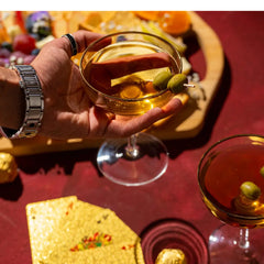 Hand holding a cocktail glass with olives, surrounded by food and another cocktail glass