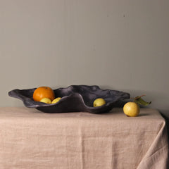 Decorative black leaf-shaped bowl with fruits on a beige tablecloth against a plain wall.