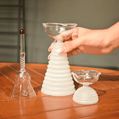 Close view of Tall glass diya with white ribbed pattern in bottom holding by a person, along with short diya and glass ringing bell 