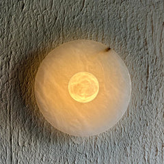 Alabaster wall sconce light fixture with a frosted outer ring and a glowing warm yellow inner circle, mounted on a textured plaster wall.