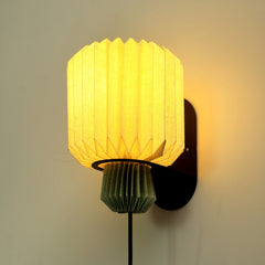 A wall-mounted lamp with a beige linen shade in a pleated design, illuminated, creating a warm light ambiance.