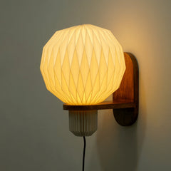 A wall-mounted lamp with a pleated, origami-inspired shade, casting a warm white glow, attached to a mango wood bracket.
