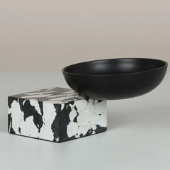 A matte black ceramic Metropia Bowl resting on a square black-and-white marbled stone pedestal against a neutral beige background.