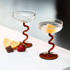 Two cocktail glasses with a swirl design on the stem. Both glasses contain a clear liquid and are placed on a white surface with a blurred background.