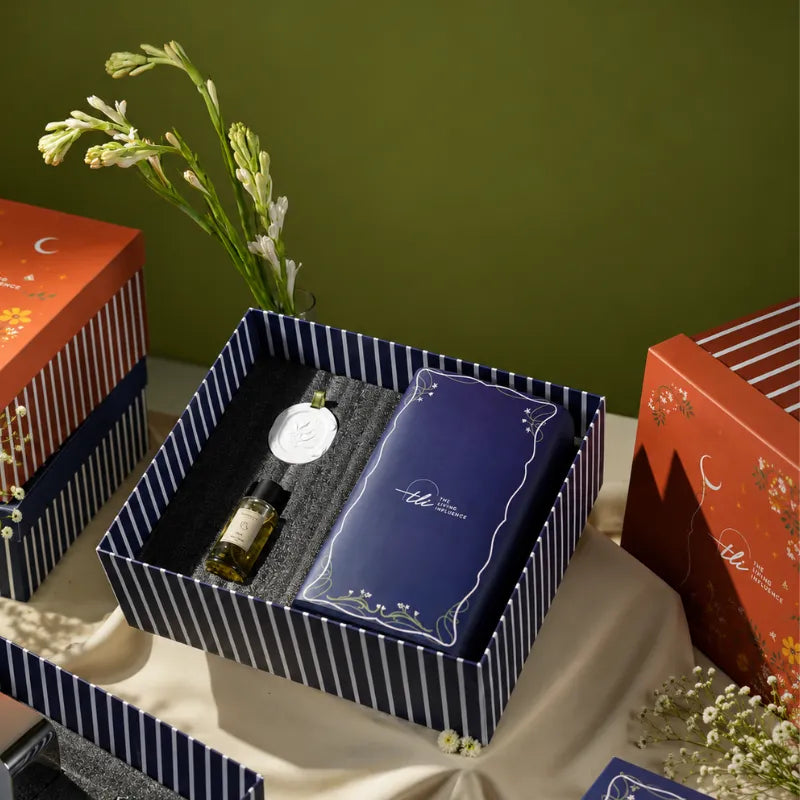 An open navy-and-white striped gift box, containing a small dark bottle of room perfume with diffusion stone and table lamp.