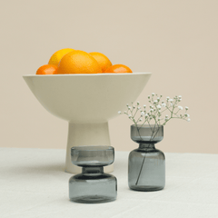 Two borosilicate glass mini vases in teal and amber colors with a small dried flower in each, placed on a table with a bowl of oranges in the background.