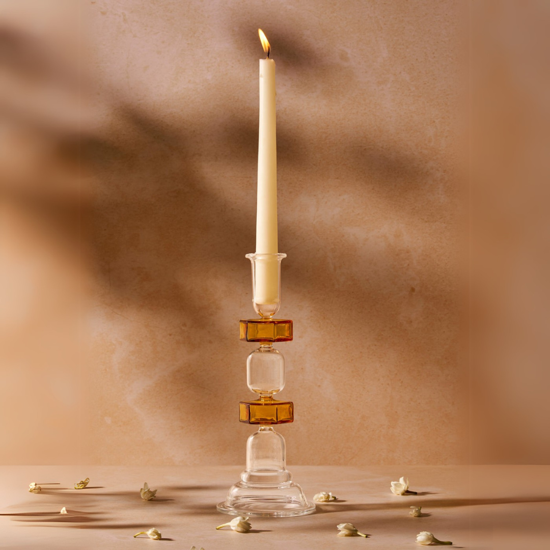 A candle with a yellow tint on a clear glass candle stand, with a tapered shape, displayed on a beige surface with some decorative elements around it.