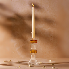 A candle with a yellow tint on a clear glass candle stand, with a tapered shape, displayed on a beige surface with some decorative elements around it.