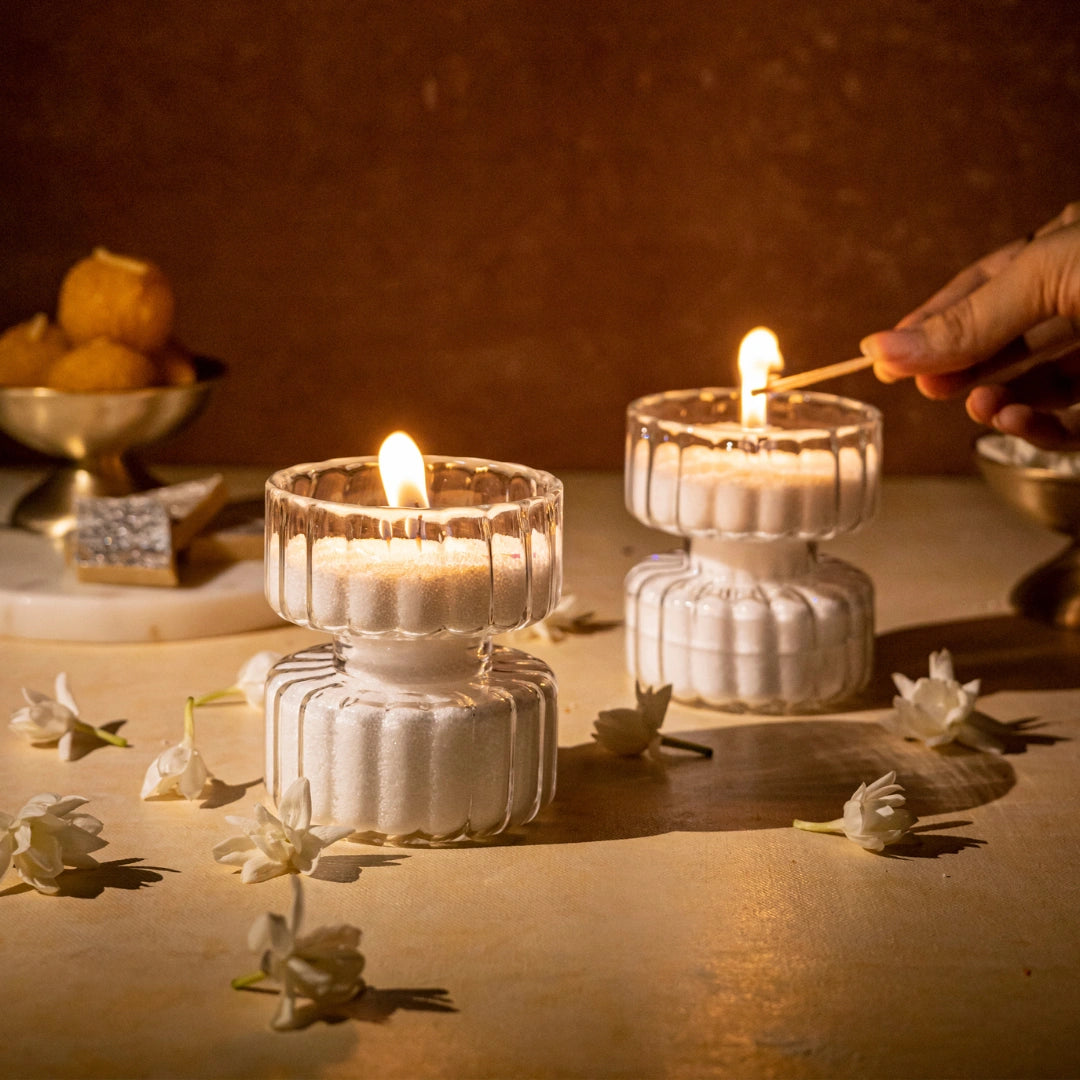 Two clear glass tea-light holders with lit candles inside, placed on a table with scattered white flower petals around.