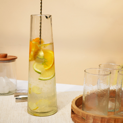 Clear glass bottle with lemon and orange slices on a textured surface with glasses and ice cubes.
