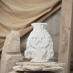 A white vase with a pixelated mosaic design, placed on a rocky surface with a textured background.