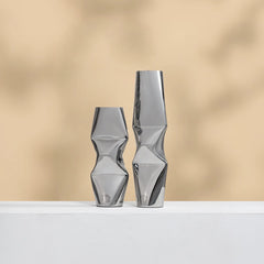 Two modern metallic silver geometric vases with a faceted, twisted design, standing on a white surface against a warm beige background with soft leaf shadows.