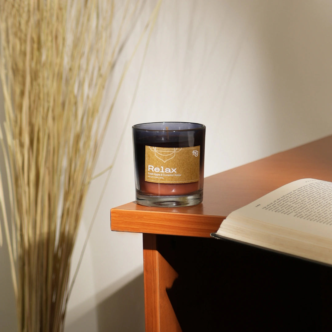 "Relax" labelled palm wax scented candle with apple and cinnamon fragrance, placed on a wooden surface, with a book and a plant in the background. 