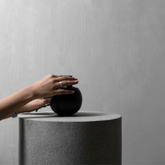 A person's hands gently touch a matte black spherical object resting on top of a textured gray cylindrical pedestal against a neutral gray wall.