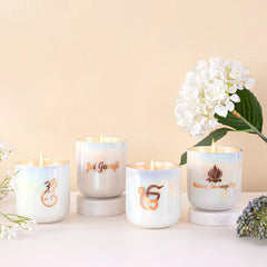 Four white U-glass candles from the Inaara range with gold-toned mantras and spiritual symbols, arranged on white marble risers with white flowers in the background.