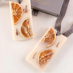 Two rectangular cream-colored Orange Cassis scented Wax Tablet - Set of 2, each featuring dried orange slices and small botanical inclusions, displayed next to gray gift box and ribbon in the background.