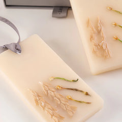 A close-up of a cream-colored rectangular Night Jasmine Wax Tablets- Set of 2 with dried wheat stalks pressed into it and a gray ribbon loop. 