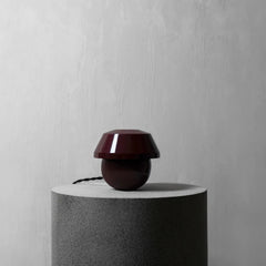 A small modern table lamp with a glossy dark burgundy dome-shaped shade and a rounded base, displayed on a textured gray cylindrical pedestal against a light gray wall.