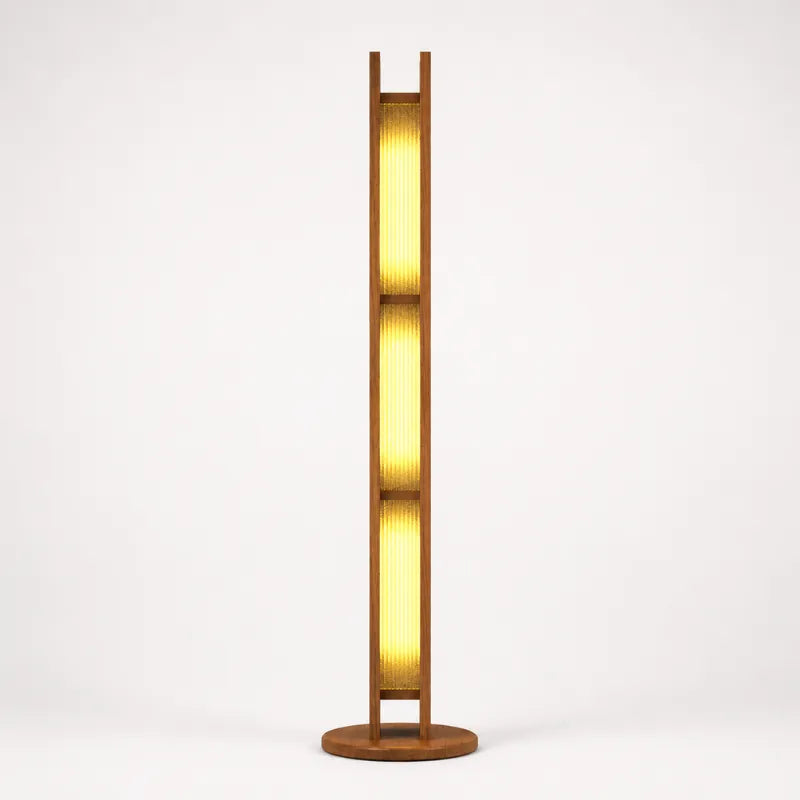 A modern minimalist floor lamp with a warm glowing vertical ribbed glass shade framed in dark wood, supported by a circular wooden base against a clean white background.
