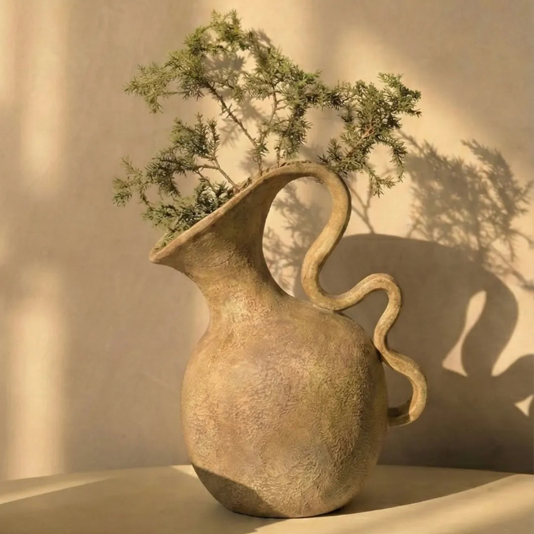 Decorative vase with a curved handle holding greenery against a beige wall.
