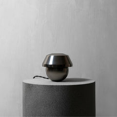 A modern dome-shaped metallic light fixture in a dark gunmetal finish, sitting on top of a cylindrical textured gray pedestal against a light gray wall.