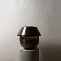 A modern table lamp with a dark bronze metallic dome shade resting on a spherical base, set on a textured gray cylindrical pedestal against a neutral gray wall.