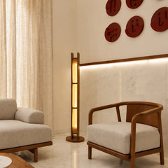 A modern interior scene featuring a tall, minimalist wooden floor lamp with integrated warm lighting, styled between a light beige armchair and a matching sofa. The background includes a cream marble accent wall with a built-in LED light strip and decorative red circular wall art.