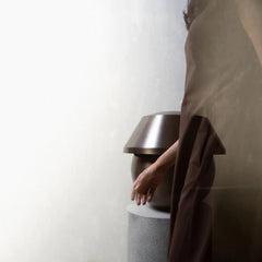 Person wearing a brown coat interacting with a modern, cylindrical object against a white wall.