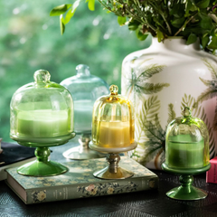 Sundance Aura Candle in yellow, grouped with different green glass cloche candle holders on pedestals. The candle holders sit on a vintage floral book in the soft-focus background. 