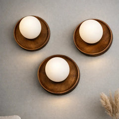 Three minimalist wall sconces with round wooden bases and glowing white globe shades, arranged on a textured gray wall with dried grass decor in the corner.