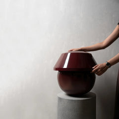 A glossy dark red spherical table lamp with a matching dome-shaped shade sits on a gray pedestal against a textured light gray wall, with a person's hands and arms adjusting the shade.