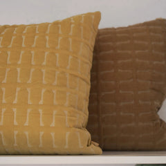 Two textured square throw pillows on a white shelf against a light gray wall. The front pillow is mustard yellow with a white geometric pattern, and the rear pillow is a warm brown with a matching embossed pattern.