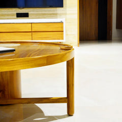 A close-up of a round wooden coffee table with a glossy finish, set in a modern living room with light-colored flooring and a wooden cabinet in the background.