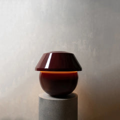 A modern table lamp with a glossy dark red dome-shaped shade and a matching rounded base, illuminated with a warm orange glow, displayed on a gray cylindrical pedestal against a textured light gray wall.