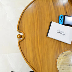 A top-down view of a round wooden coffee table with a decorative scalloped edge and a small round marble coaster. A white Studio Lakdoo box and a blue Nintendo Switch are placed on the table.