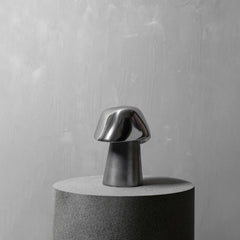 A modern mushroom-shaped metallic table lamp with a reflective silver cap and a tapered silver stem, displayed on a cylindrical gray pedestal against a textured light gray wall.