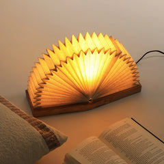 A linen book lamp with linen shade & mango wood base illuminated, placed next to an open book.