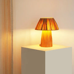 A Petite Table Lamp with a tiered, woven design, emitting a warm glow, placed on a white surface in a dimly lit room.