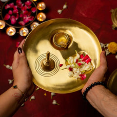 Gold Pratah Pooja Brass Thali & Bell with flowers and a candle held by two hands on a red background