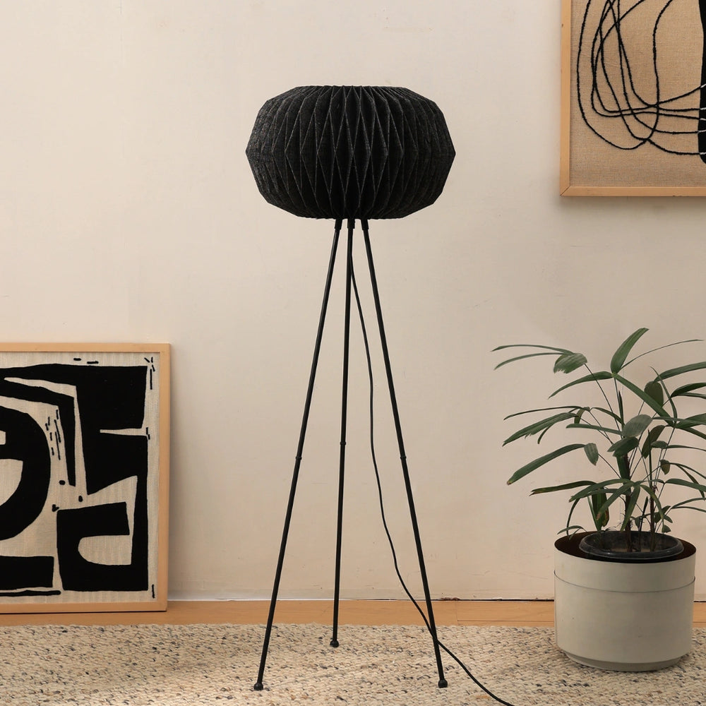 A modern origami-inspired floor lamp with a black linen shade on a tripod stand, standing in a room with decorative plants and wall art.